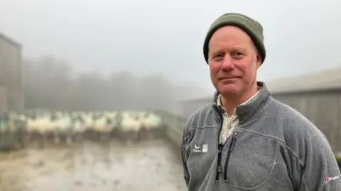 Farmer Neil Cole wears a grey fleece over a pale checked shirt, with his flock in the background 