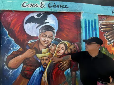 A man in a black hat and shirt points to a colourful mural painted on a wall showing a man carrying 4 people - three of them sleeping and one with his eyes open wearing a cowboy hat. The many carrying the farmworkers is meant to be Cesar Chavez and he is carrying a red flag. 