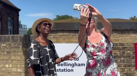 Two women in dresses and sunglasses smile for a selfie in front of the new station sign.