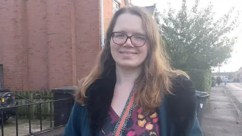 Katherine Coffee smiling at the camera. She has long red hair, wears brown-framed glasses, a velvet suede fur coat, and a purple, orange, and black floral-print blouse. She is standing on a sidewalk with containers behind her.