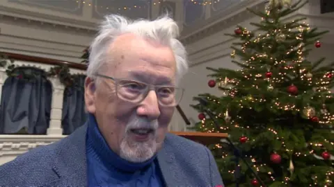 Grenville Jones stands beside a Christmas tree. He has white hair and is wearing a blue suit jacket and a knitted jumper.