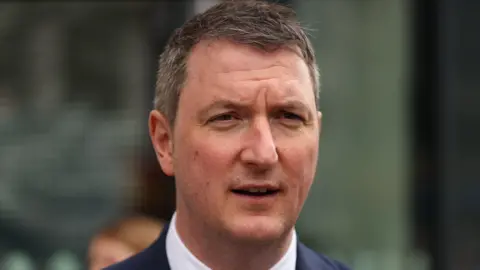 PA Media John Finucane is standing outside wearing a navy suit and white shirt. He has short brown hair with white hairs on the side. He is mid-speech. 