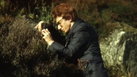 Rick Cornwall Rick is wearing a black coat, has brown/ginger hair and glasses and is looking at coastal heather in 1976