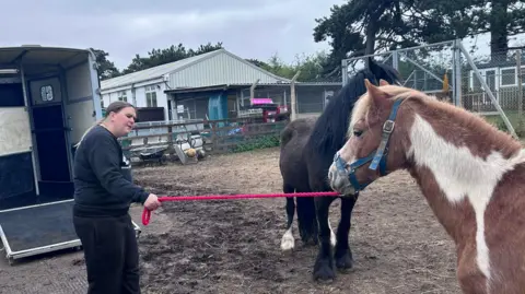 BBC/Kate Bradbrook A woman dressed in black stands in front of a horsebox holding a brown and white pony by a red lead rope. She is taking the pony to its new home at the charity's second site.