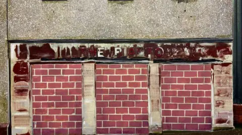 A painting by Narbi Price called "If you want to change things you cannit change them from the floor". It looks like a closed down business and there is an illegible sign painted above what look like bricked up doorways or windows.