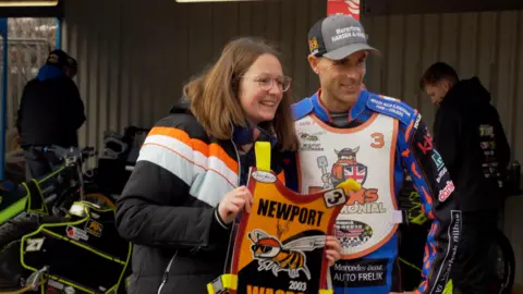 Jonny Michel/BBC Iversen is standing in his blue racing jacket with lots of branding stuck on. He is posing for a photo, standing beside a woman with shoulder length hair and glasses, who is holding a Newport Wasps racing bib