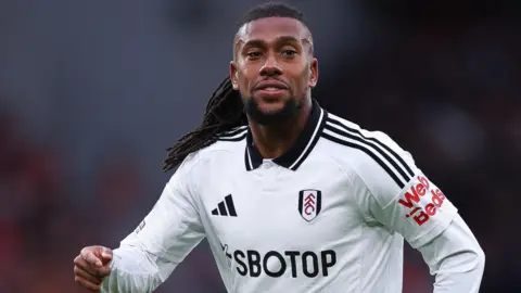 Alex Iwobi, wearing a white Fulham shirt, looks to the right of screen as black dreadlocks are seen over his left shoulder and he raises his right arm to chest height