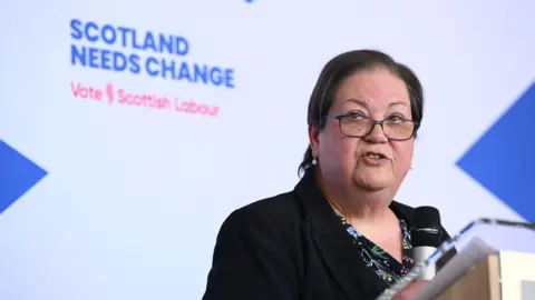 PA Media Jackie Baillie standing at a podium in front of a large saltire background with a Scottish Labour election message on it.