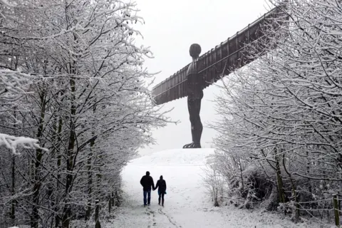 Owen Humphreys/PA Media A snow-topped Angel of the North sculpture stands atop the hill. A couple of people, holding hands, are walking up towards it and leaving snowy footprints behind them. Their path is bordered by snow-topped trees.