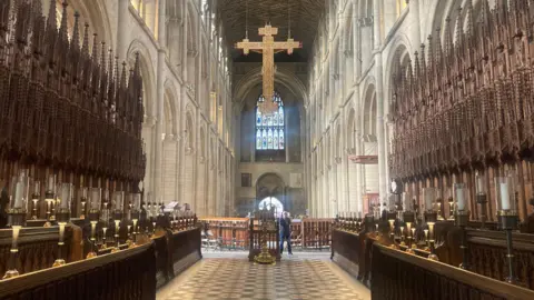 Mousumi Bakshi/BBC The nave of Peterborough Cathedral, with a big golden cross hanging from the ceiling.