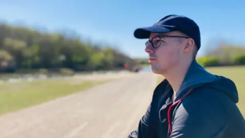 Jake is sitting alone on a bench in a park with a path beside him. 
He is casually dressed and wearing a dark cap and glasses. He is looking into the distance with a thoughtful expression.       
