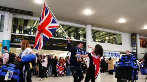 PA Media A man, Matt Weston, is about to be hugged by a woman wearing a union jack flag as a cape. He is carrying a large union flag and wearing gold medals around his neck. They are stood in the middle of an airport arrivals lounge.