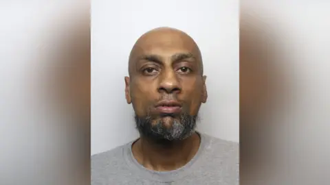 A bald man with a greying beard and grey t-shirt, pictured in a police mugshot. 