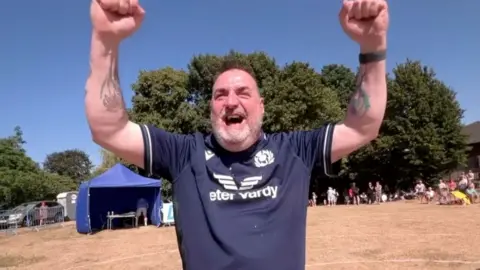 A man in a blue sports shirt raising his arms in the air in a field