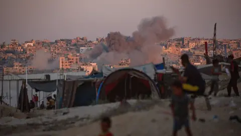 Reuters - Rook steeg op na een Israëlische luchtaanval, terwijl ontheemde Palestijnen naast tenten zaten in Khan Yunis in de zuidelijke Gazastrook (13 augustus 2024)