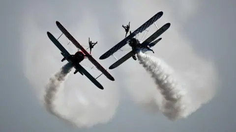 Getty Images Two planes flying in the sky with smoke trails coming off the back. On the top of each plane is an acrobat