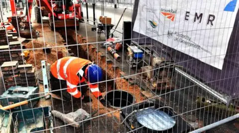EMR a man in an orange high-viz jacket and blue helment crouches in a trench lined with sand.