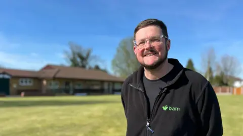 Steve Murphy stands in front of the clubhouse at Water Orton Cricket Club, he is wearing a black fleece and t-shirt and has short brown hair and wears glasses. The grass has been freshly mown and the sky is a vivid blue.
