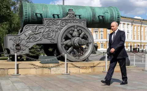 Getty Images Putin walks past a Tsar Cannon monument in Moscow. He is wearing a dark suit, blue tie and white shirt. The cannon is very ornate and situated on a pedestal.