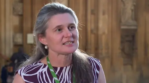 A grey-haired woman in a purple and white zebra-style patterned dress with a green lanyard on. She is in front of gothic-style limestone wall.