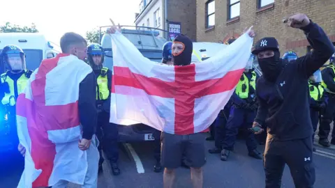 PA Media Three protesters standing in front of a line of police officers. One has an England flag draped over his back. The others both have their faces covered and one is holding an England flag.