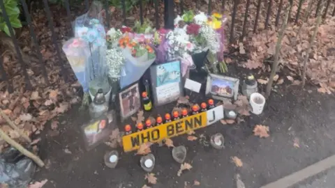 Supplied A collection of cards, flowers, candles and a personalised registration plate sat against a metal fence. Autumn leaves are scattered around.