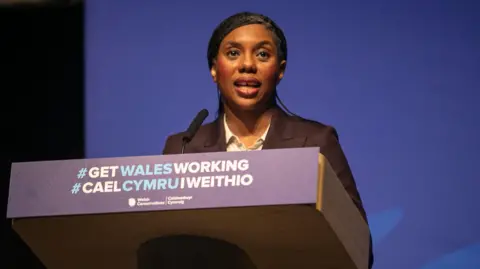 Welsh Conservatives Kemi Badenoch stood at a lectern that reads "#Get Wales Working" in Welsh and English.