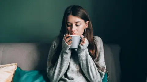 A young woman with a grey jumper on sits on a sofa and is holding a hot drink.