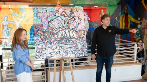 Trivandi A painting in the middle of the photograph, with a ginger woman on the left hand side in a blue shirt and white trousers and her arms crossed. On the right hand side of the painting is a man in a black shirt and dark blue jeans looking over to the right side of him and pointing. 