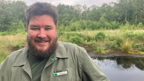 Dr Paul Thomas with brown hair and beard wearing khaki green shirt and t-shirt with his name badge on standing in Risley Moss in Birchwood with a stream of water behind him and trees in the background. He is smiling.