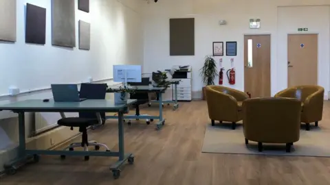 Salt Arts CIC Inside the new facility. The image shows a white walled office room with three class desks. There is a sitting area to the side of the desks with three yellow/brown leather chairs.