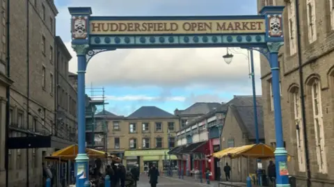 Olivia Courtney-Ashton/BBC Image shows street scene with market stalls. There is a large blue archway which sign posts the start of the open market. 