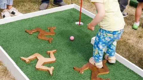 Martin Parr/Magnum Photos A toddler plays crazy golf. They're standing on a putting green, with artificial grass, which has wooden triskelion's. They are wearing flowery crocs, and blue flowery jeans with a yellow t-shirt.