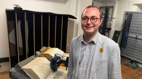 A man in a grey suit with glasses stands in frount of technology which is scanning ancient Bibles.