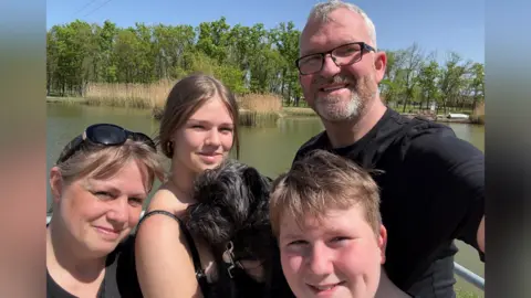 Bruce Lait takes a selfie in front of a lake with his daughter, his son and his wife. They are all smiling. His daughter is holding a small black dog.
