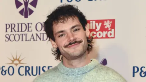 A man with a brown mullet and moustache wearing a light green knitted jumper. He is standing in front of a white backdrop.