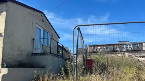 A cream concrete bungalow with black stains on the wall in front of some wasteland which is surrounded by metal fencing