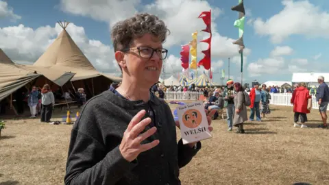 An author with short grey hair and a dark grey cardigan is holding a small book titled Teach Your Dog Welsh. In the background there is dry yellowish grass, a neutral colored tan tent and bright flags billowing in the wind in various colors like red, yellow and light green. 