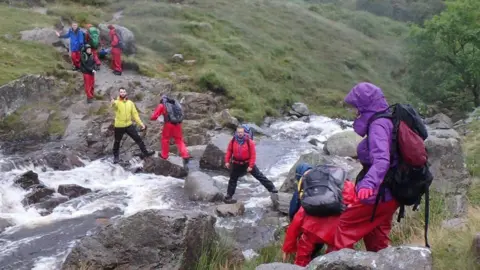 Sarah Donkin A group of youngsters cross a river. They are wearing outdoor clothing and carrying backpacks.