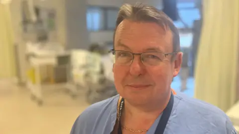 Dr Greg Fletcher wearing blue theatre scrubs standing in the intensive care unit at the Royal Derby Hospital
