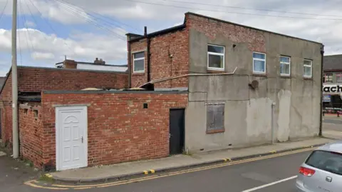 Google Street view of 8 Stockton Terrace. It is a two-storey building of red brick and peeling cracked gray render. A white door is cut into the brick wall and upstairs there are four windows.