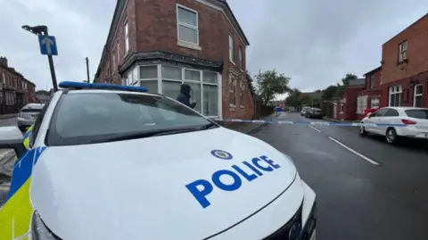 A police car in the foreground of the image, which shows a road on the left and the right. A white car and other vehicles further away are behind police tape on the right.