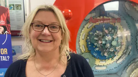 Nicola Masters smiles at the camera. She has shoulder-length blonde hair, black rimmed glasses and is wearing a navy blue cardigan. She stands in front of an Aardman display at the hospital.