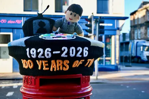 Charlie McGinn A knitted post box topper depicting John Logie Baird's first public demonstration of a working, mechanical 'televisor' system and the words '1926-2026: 100 years of TV'. There are buildings in the background, and a lorry.