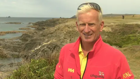 Martin White is standing at the top of a grassy cliff looking over the rocky beach. He is smiling towards the camera and is wearing a red fleecy jacket. He has white hair and has a sunglasses on his head.