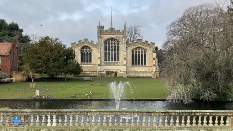 Amy Holmes/BBC A church in the background with a lake and a fountain in front. There are ducks on the grass behind the lake which has some railings around it and trees.