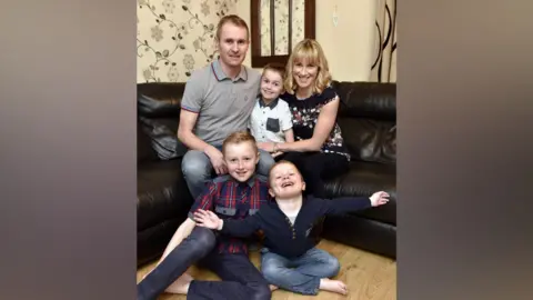 David Hollins Tony and Sure at home with their children Daniel, 9, Adam, 7, and Jack, 4. They are sitting on a corner sofa and are all smiling at the camera.