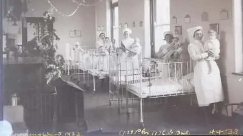 South West Heritage Trust Nurses dressed in uniforms with aprons and headdresses hold babies beside their hospital beds in Bowring ward in the Royal Devon and Exeter Hospital. There is a Christmas tree in the background.

