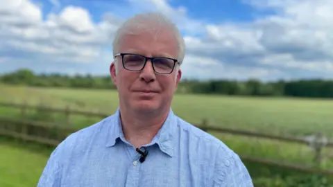 BBC Man in his sixties wearing a blue shirt in front of open countryside