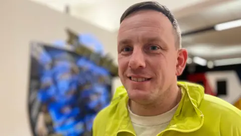 Emma Baugh/BBC Nathan, a man, with short light brown hair, wearing a yellow zipped-jacket and a white shirt, smiling for the camera.